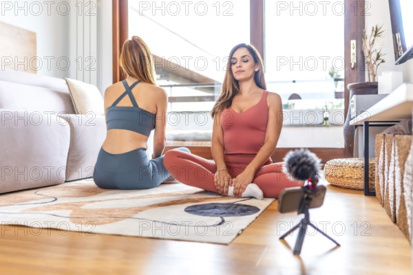 Two women recording an online yoga and meditation class in a cozy living room, embracing wellness