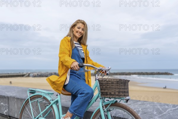 Smiling woman with yellow raincoat sitting on a light blue bicycle by the sea enjoying freedom and