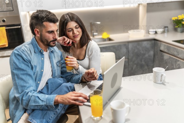 Young couple sitting at kitchen table, drinking orange juice and using laptop, enjoying their time
