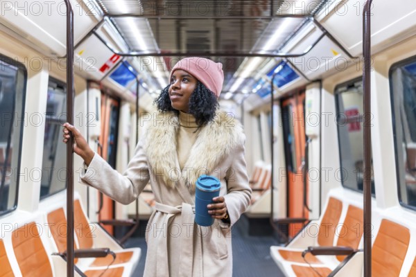 Young black woman wearing winter coat and pink woolen hat holding handrail and reusable coffee cup