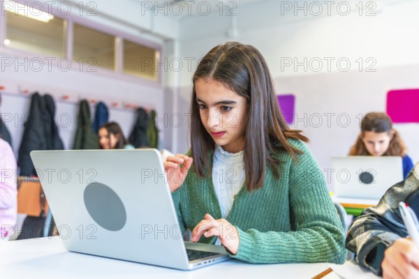 Schoolgirl concentrating on her laptop during a lesson, surrounded by classmates in a modern