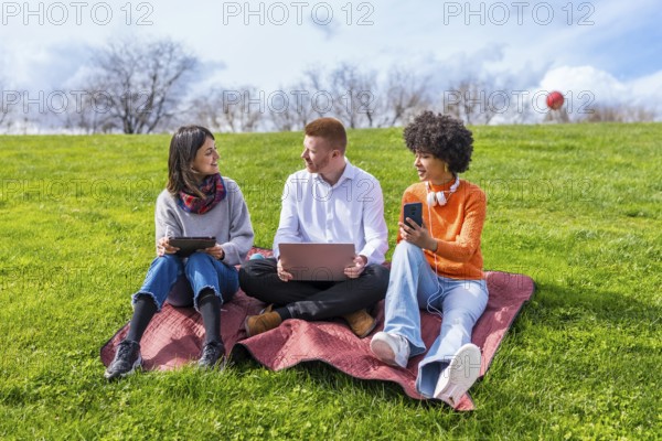 Three university students using electronic devices while sitting on a blanket in a park, enjoying a