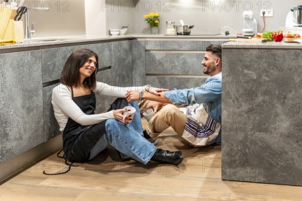 Two chefs are sitting on the floor of their modern kitchen, enjoying a coffee break and chatting
