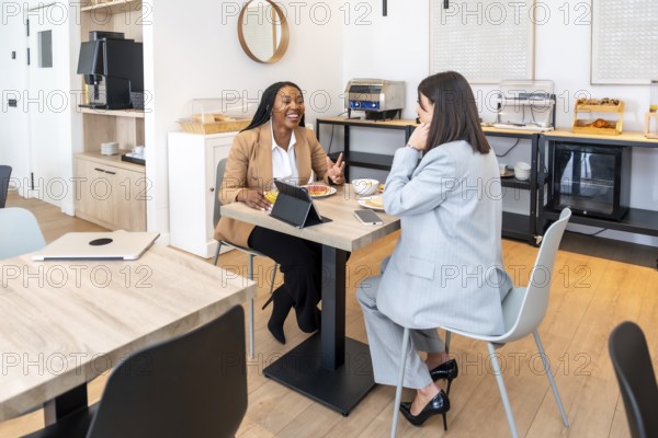 Two businesswomen enjoying breakfast and discussing work during a business trip, utilizing a tablet