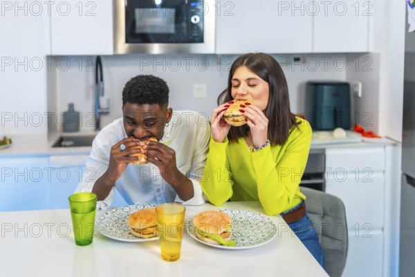 Young multiethnic couple eating homemade burgers in their modern kitchen, enjoying a casual meal