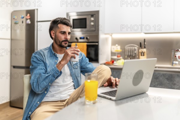 Man savoring a cup of coffee while working remotely on a laptop in a stylish, modern kitchen,