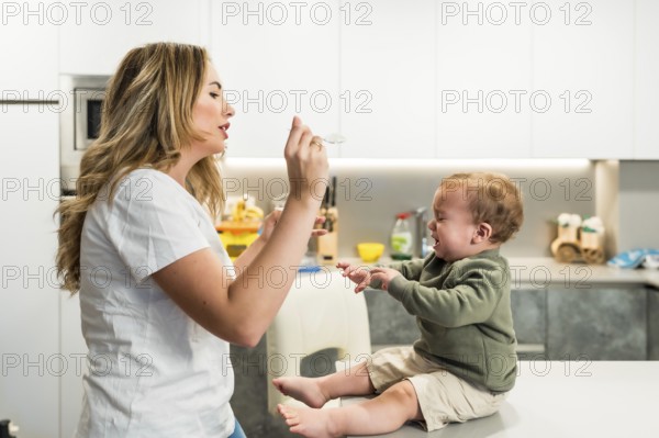 Young mother struggling to feed her crying baby boy while sitting on the kitchen counter at home,
