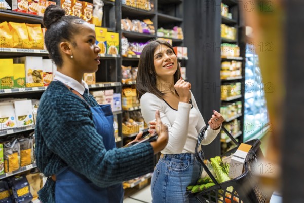 Saleswoman using tablet, advising customer holding shopping basket, choosing products on shelves in