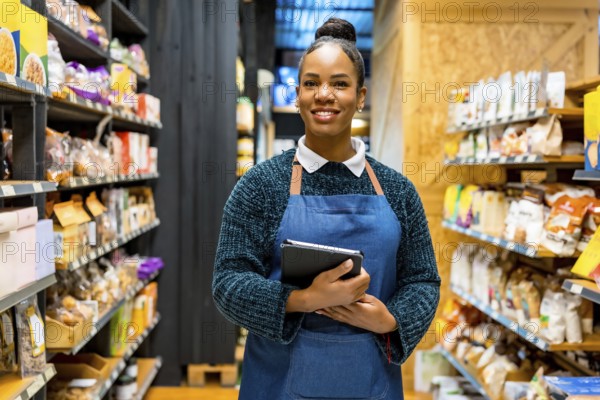 Confident saleswoman in an apron, smiling while holding a digital tablet and working among shelves