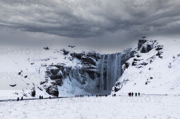 Tourists at Skogafoss waterfall, snowy and icy rock face, dark clouds above, Sudurland, Iceland,
