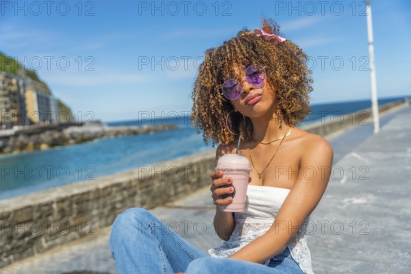 Cool latin young woman with curly hair in summer clothes drinking a shake outdoors next to the sea