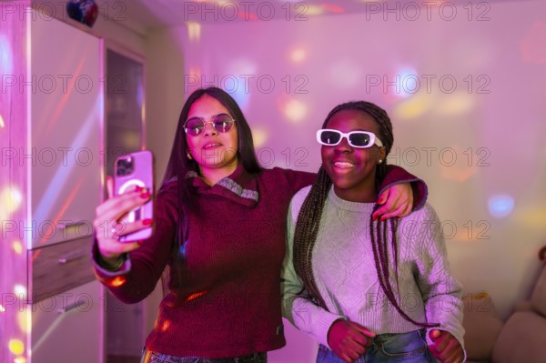 Two happy teenage girls with sunglasses taking a selfie with smartphone at a home party with disco