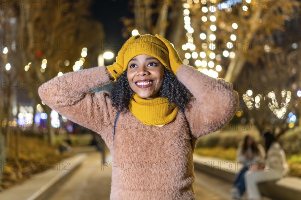 Smiling young woman wearing winter clothes adjusting her yellow wool cap under christmas lights in
