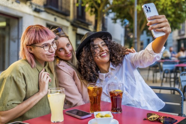 Happy female friends taking selfie together sitting on a sidewalk cafeteria drinking soda and