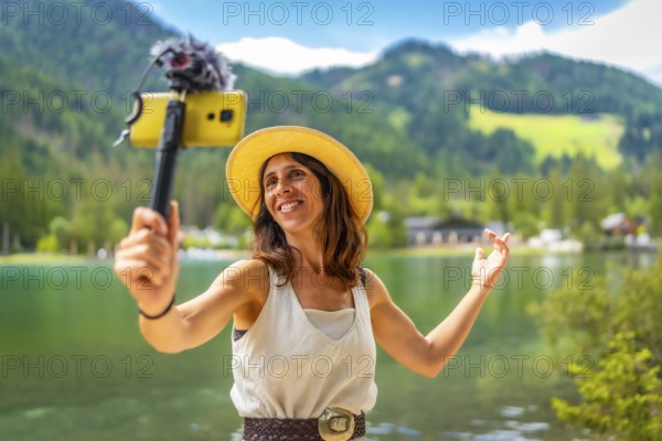 Smiling woman wearing straw hat vlogging with smartphone and external microphone at lake dobbiaco