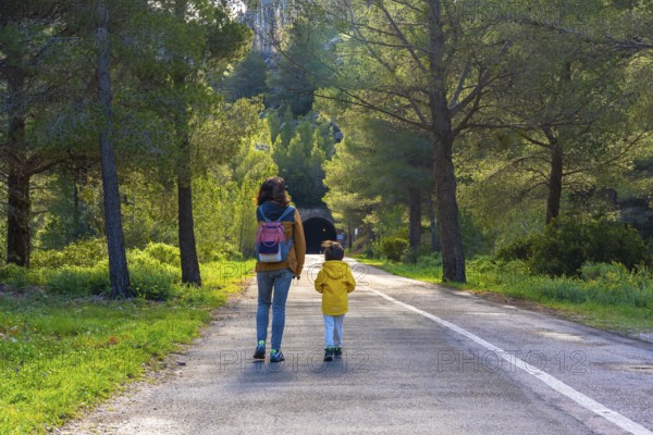 Mother and child enjoying a leisurely walk on a paved road amidst lush greenery and pine trees near