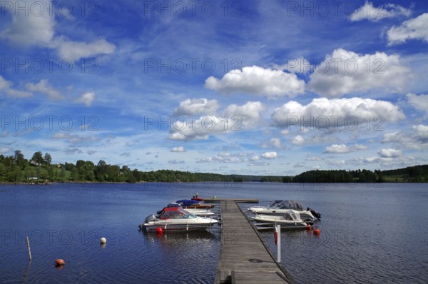 Lake with a wooden jetty and boats moored on it under a blue sky with white clouds, Stora Le,