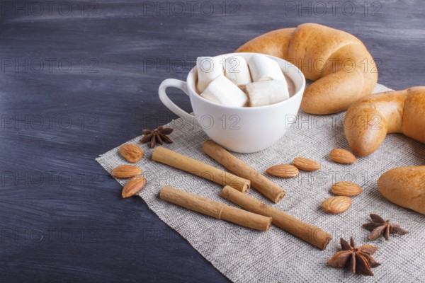 A cup of hot chocolate with marshmallow, croissants and spices on linen napkin and black wooden