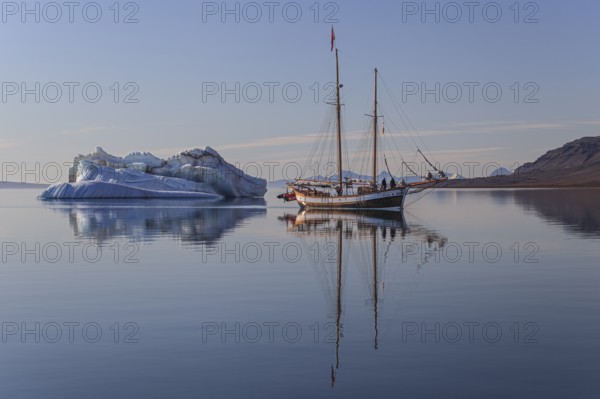 Traditional sailing ship reflected in fjord next to iceberg, sunny, Ittoqqortoormiit, Scoresby