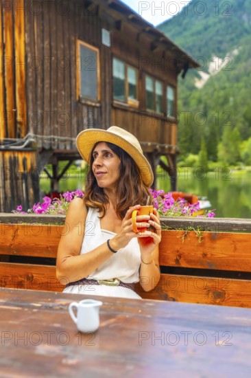 Woman wearing straw hat holding a mug and enjoying a hot beverage at a wooden table with lake