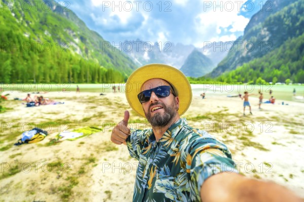 Tourist wearing sunglasses and straw hat gesturing thumbs up while taking a selfie at lago di