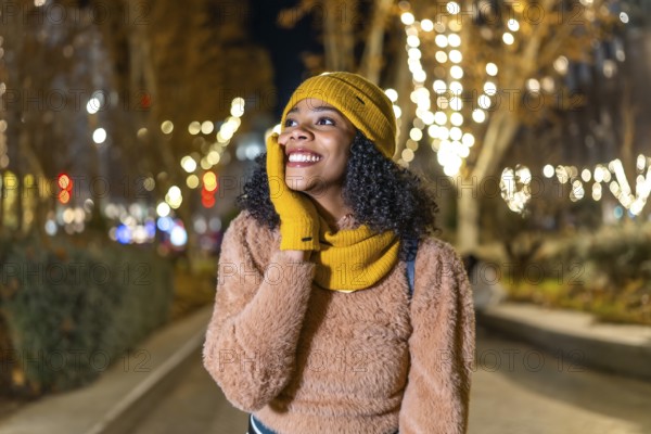 Smiling young woman wearing winter clothes touching her face while enjoying christmas lights in a