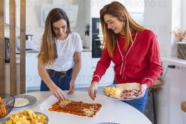 Two women are happily adding cheese to a pizza base while preparing a meal together in a modern