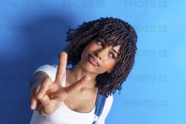 Young woman with curly hair making a victory sign with her fingers against a vibrant blue