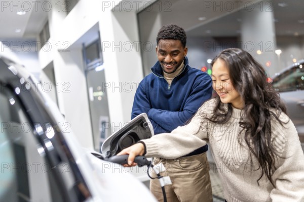 Happy multiethnic couple charging their new electric car at a dealership showroom, embracing a