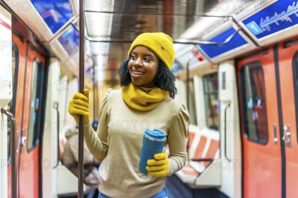 Smiling woman wearing yellow woolen hat and gloves holding a reusable mug while standing inside a