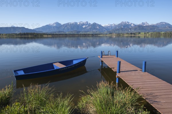 A lake with calm water and blue sky, a boat and a jetty in front of an alpine backdrop, Hopfen am
