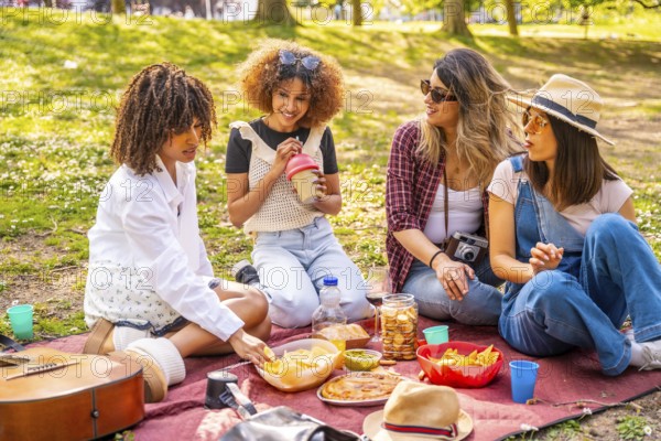 Four female friends are sitting on a blanket, eating snacks and drinking beverages during a picnic