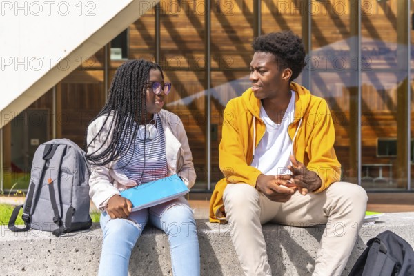 Two young African american students chatting relaxed outside the university sitting together under