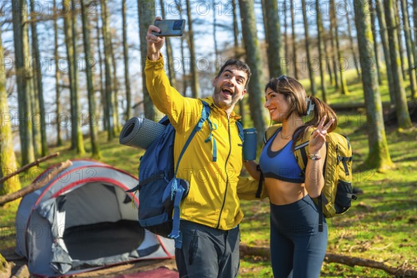 Happy couple of hikers taking a selfie with smartphone in a forest next to their tent during a