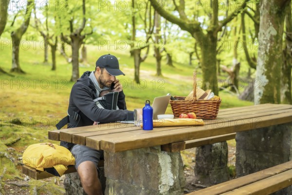A young Caucasian father teleworking sitting at a picnic table with his computer and talking on the