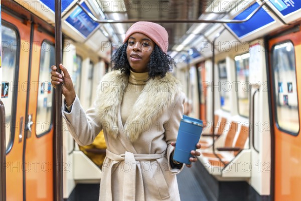 Stylish young black woman wearing winter coat and pink beanie holding reusable coffee cup while