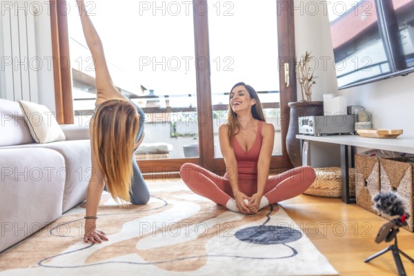 Two female friends practicing yoga together at home, following an online class on a device off