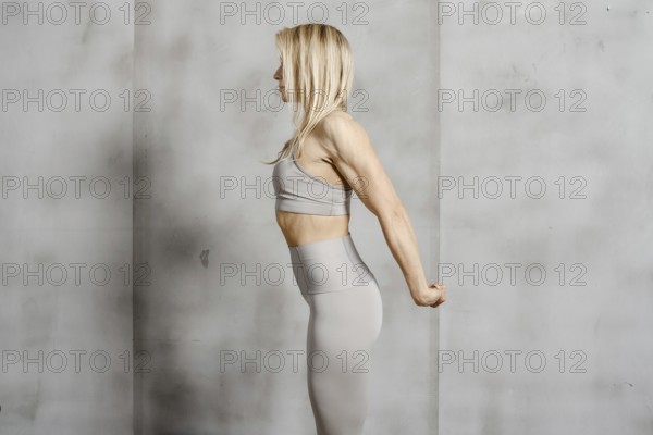 An athletically built woman stands in profile against the background of a concrete wall and