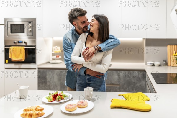 Young loving couple hugging and laughing in contemporary kitchen, with croissants, strawberries,