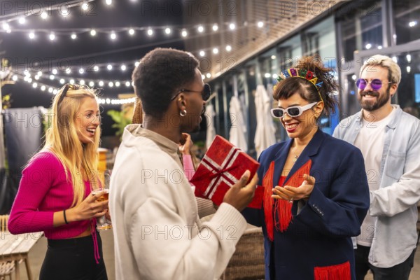 Multi-ethic friends giving a gift box to a woman celebrating birthday in the terrace of a modern