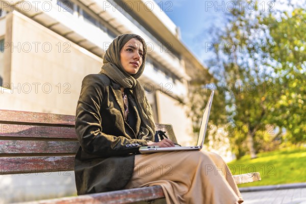 Side low angle view of a muslim businesswoman with headscarf working on remote sitting on park
