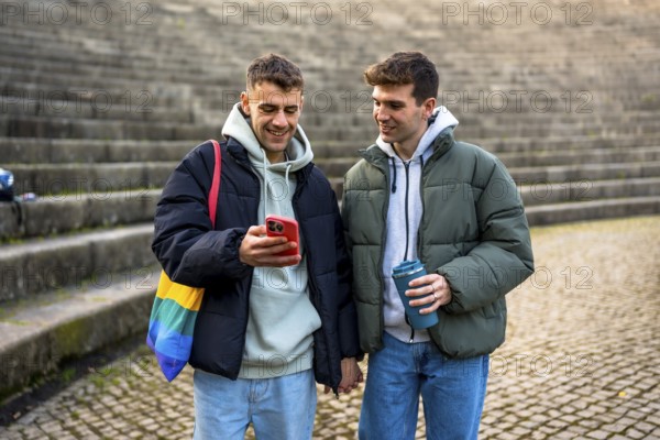 Happy gay couple holding hands, looking at smartphone and enjoying a reusable cup of coffee