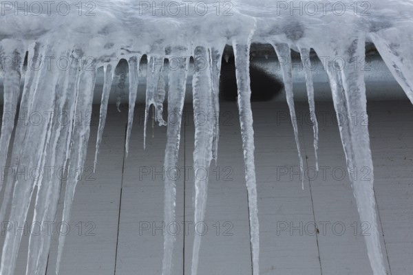 Long icicles and thick heavy ice hanging from roof edge of grey wood plank cladded building in