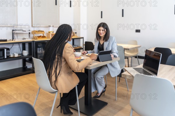Two businesswomen enjoying breakfast and discussing work using a digital tablet in a modern hotel