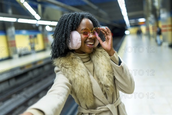 Smiling young woman wearing earmuffs and stylish glasses taking a selfie while waiting for the