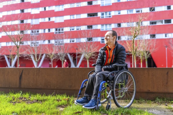 Young man sitting in wheelchair enjoying a sunny day in a modern urban park with a red building in