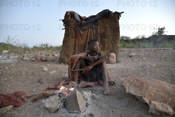 Young shepherdess from the Himba people sitting at a campfire in front of her hut, Ombombo,
