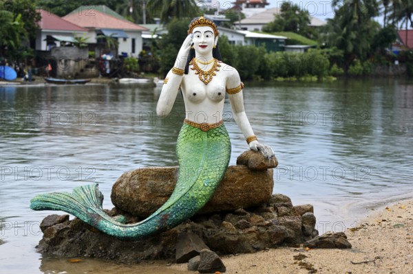 Greeting mermaid statue with white upper body and green tail fin sits on a stone on the shore,
