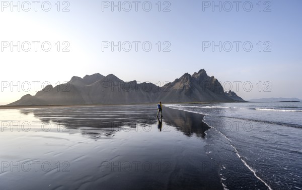 Tourist at black lava beach, sandy beach and sea, mountains Klifatindur reflected in the water,