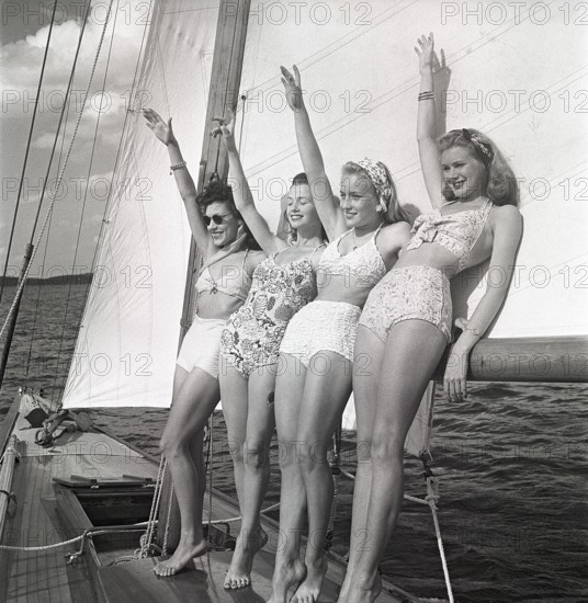 1940s sailing boat. Four young women are standing and leaning against the sails of the boat waiving